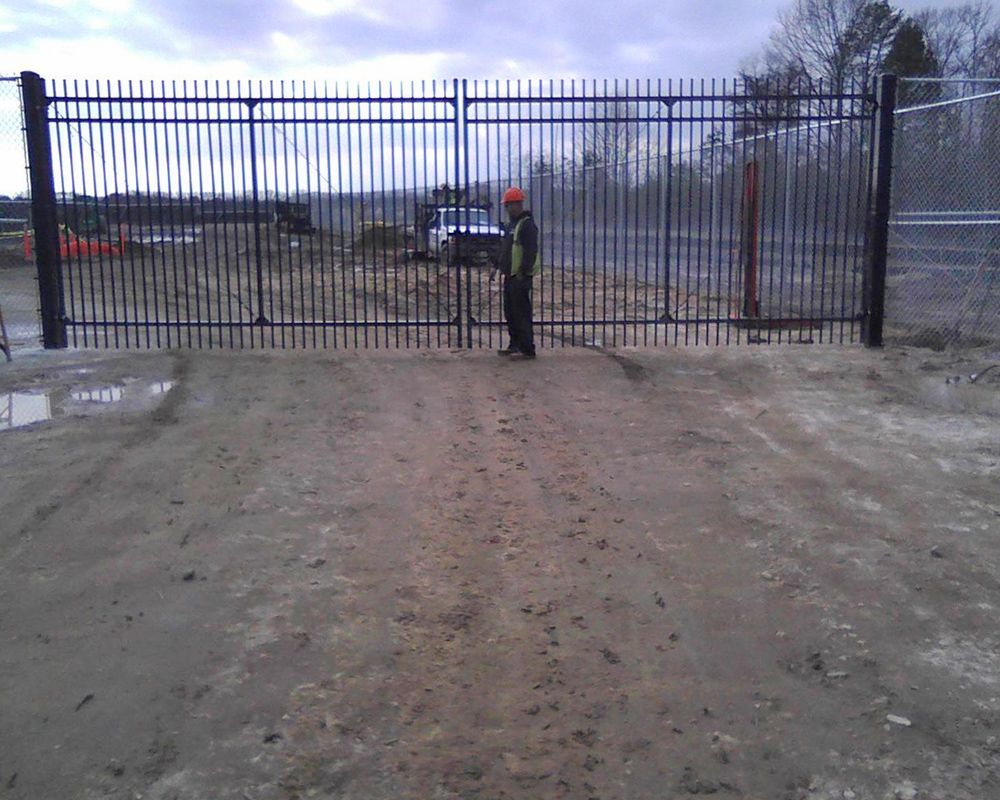 Person standing by a tall, black metal gate on a dirt road, likely construction site.