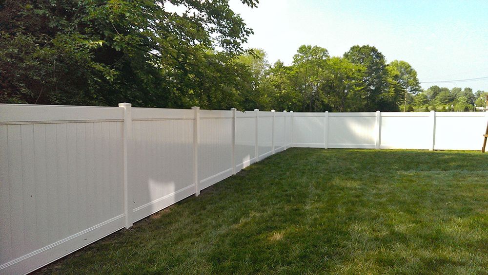 White vinyl fence encloses a green lawn, trees in the background under a bright sky.