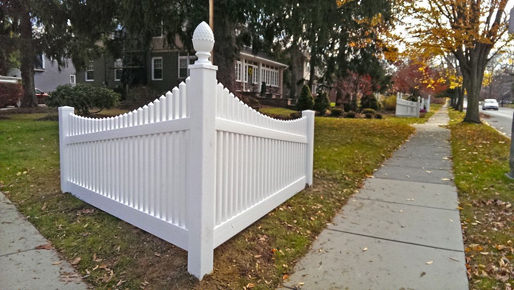 White picket fence on a grassy lawn next to a sidewalk and a street with houses in the background.