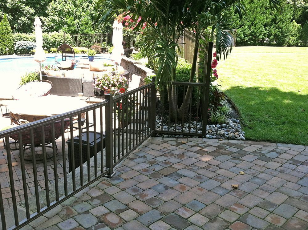 Patio with brick pavers, metal railing, palm tree, and pool in the background. Sunny outdoor setting.