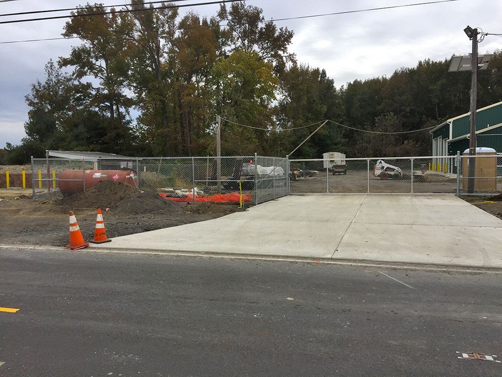 Concrete driveway entrance behind a chain-link fence, construction site with trees in background.