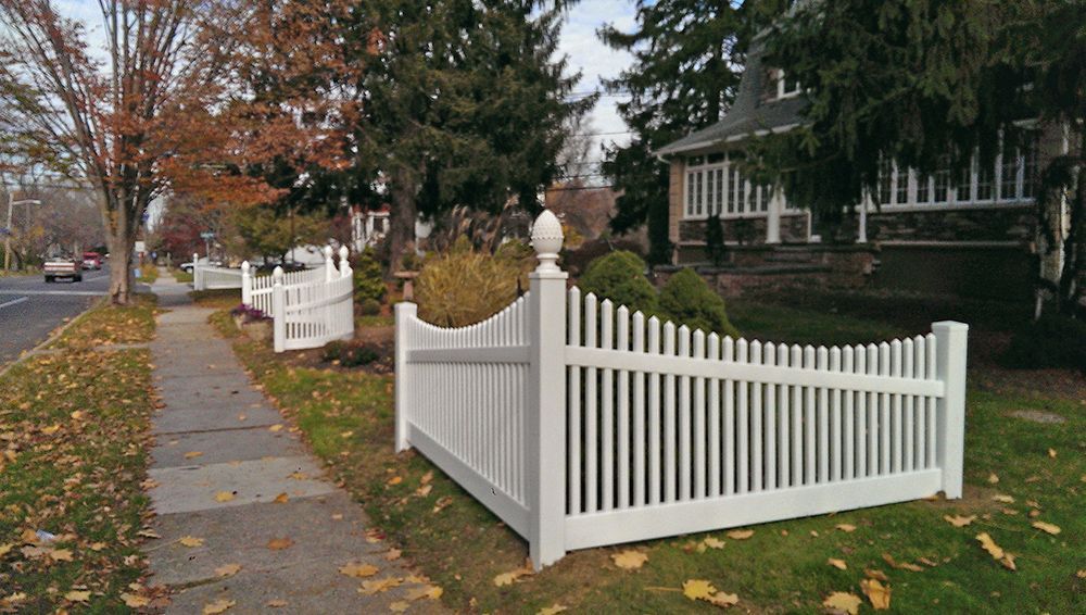 White picket fence bordering a sidewalk and lawn, trees, and houses along a street.