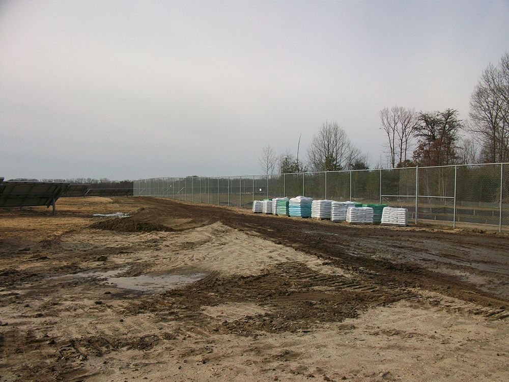 Chain-link fence borders a muddy construction site with piles of building materials and bare trees under an overcast sky.