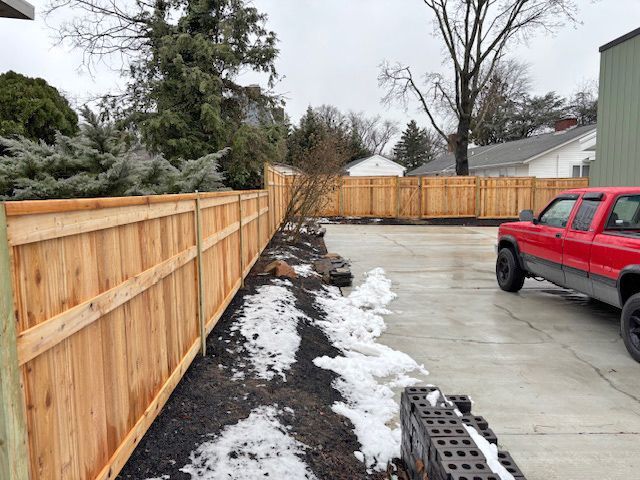Wooden fence along a driveway with a red truck parked on the right and snowy ground in the foreground.