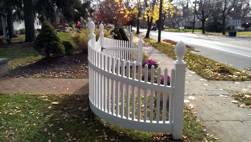 White picket fence curving along a sidewalk with fall foliage and a road.