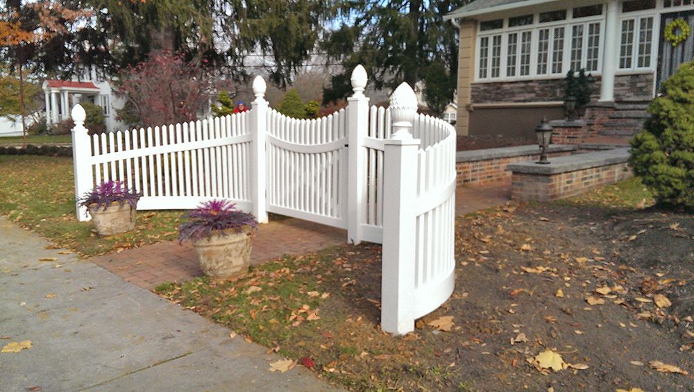 White picket fence curving along a brick pathway, flower pots, and a house in the background.