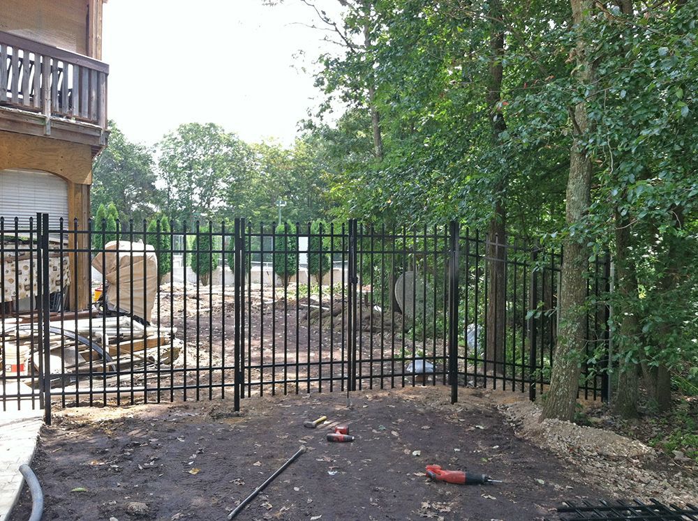 Black metal fence surrounds a backyard with trees and a building on a sunny day.