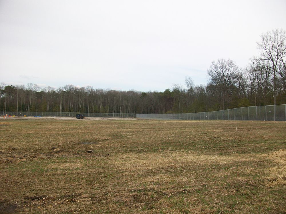 Grassy field with a tall, chain-link fence and a line of trees in the background under a cloudy sky.
