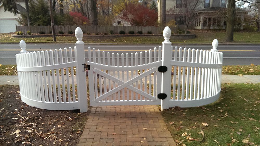 White picket fence with a gate, curving across a brick pathway in front of a house.