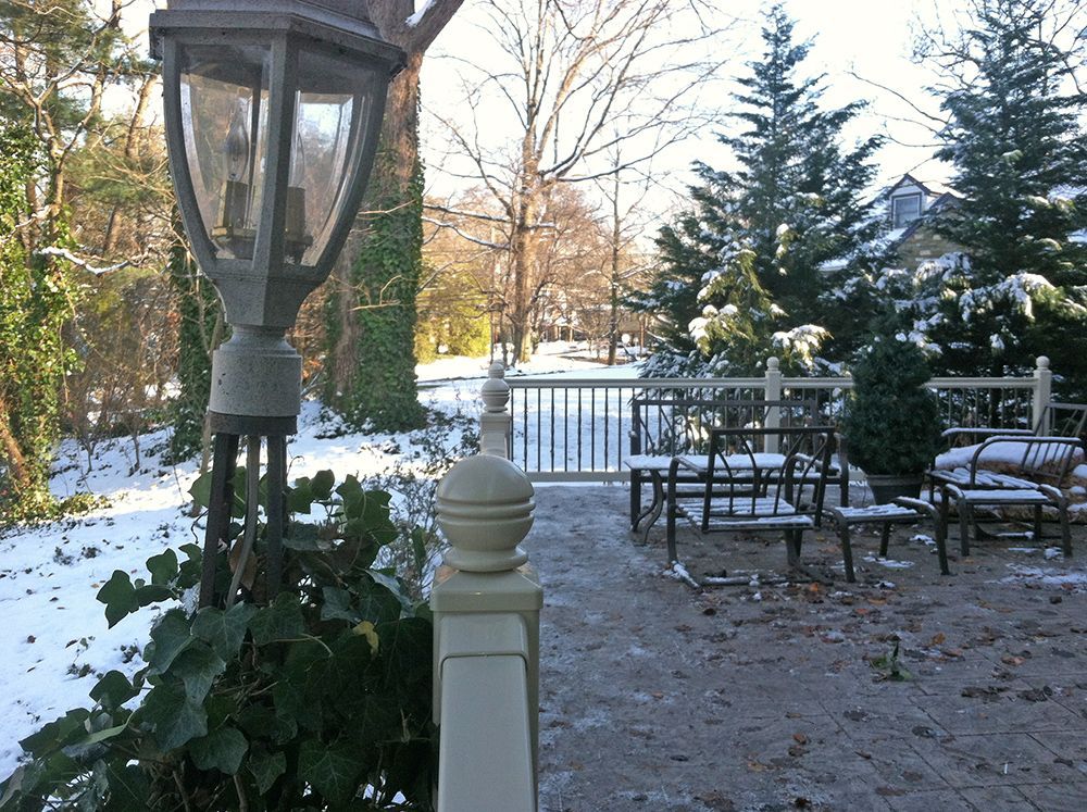 Snow-covered patio with chairs, a light post, and evergreens. Winter scene with a wooden fence leading to a park.