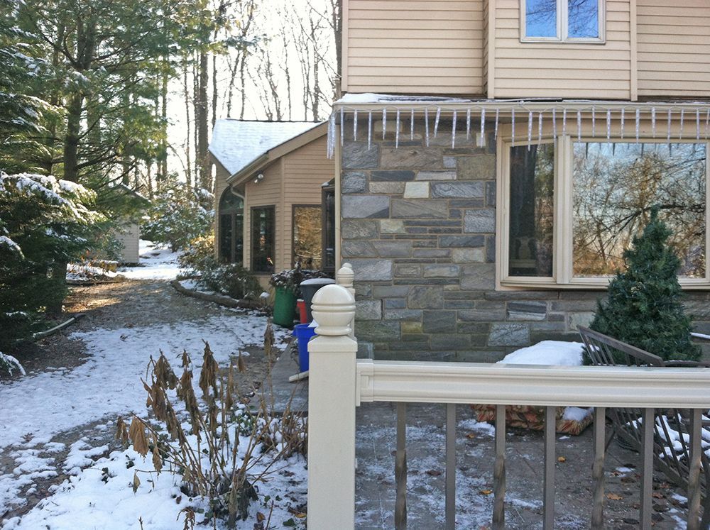 Snowy backyard with stone-faced house, icicles, and walkway leading to another building.