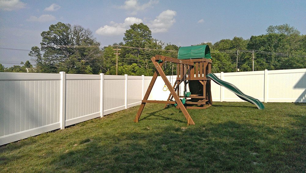 A backyard with a white fence, green grass, and a wooden playset with a slide and swings.