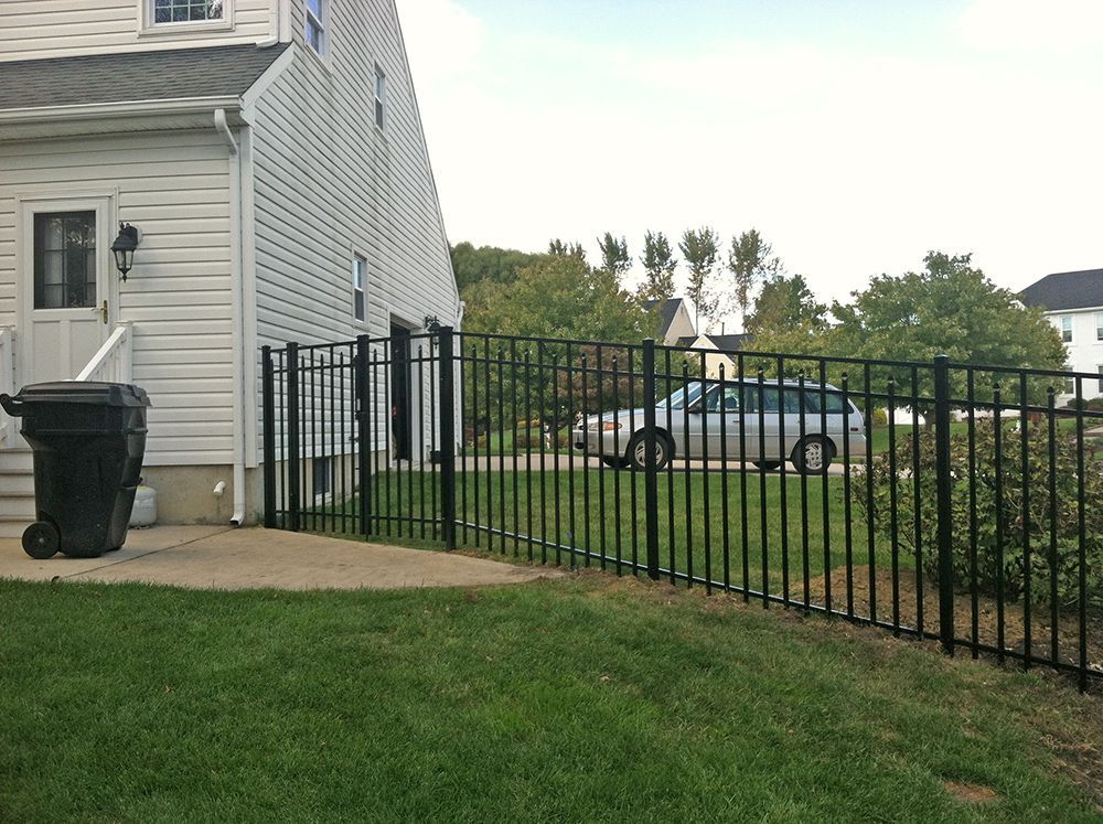 Black metal fence next to a two-story beige house with a black trash can on a concrete patio.