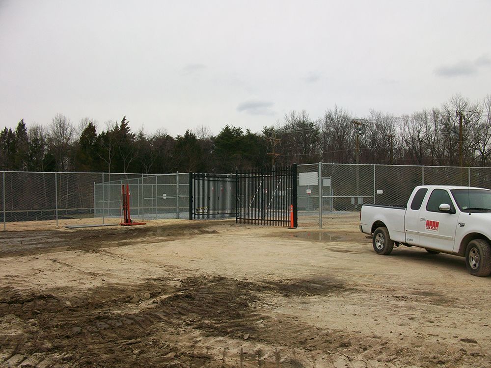Fenced industrial site with gate, truck parked on dirt lot, trees in background on cloudy day.
