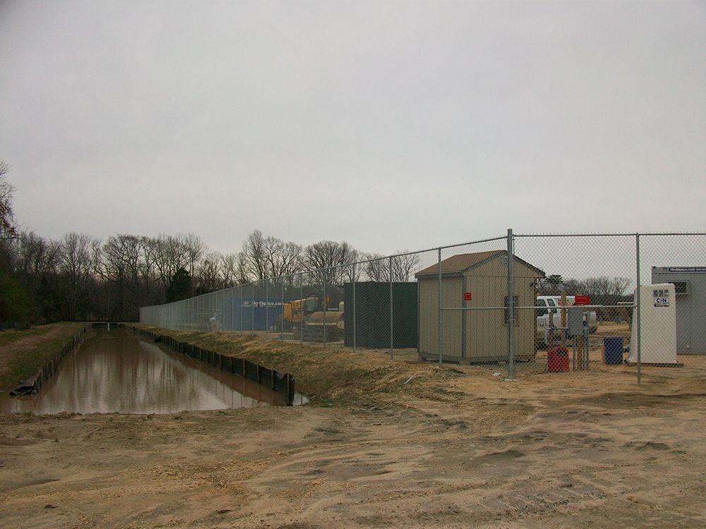 A muddy area next to a water channel, behind a chain link fence with a small shed.