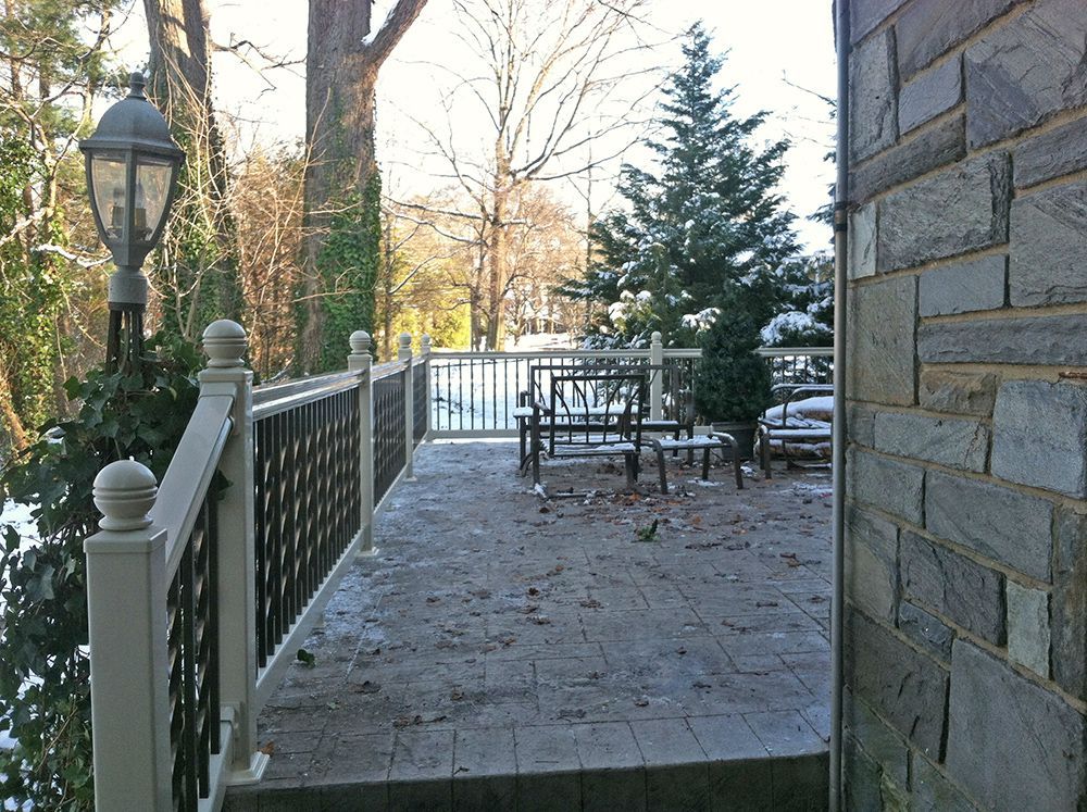 Outdoor patio with decorative railing, stone wall, and snow-covered furniture. Trees and a lamppost are visible.