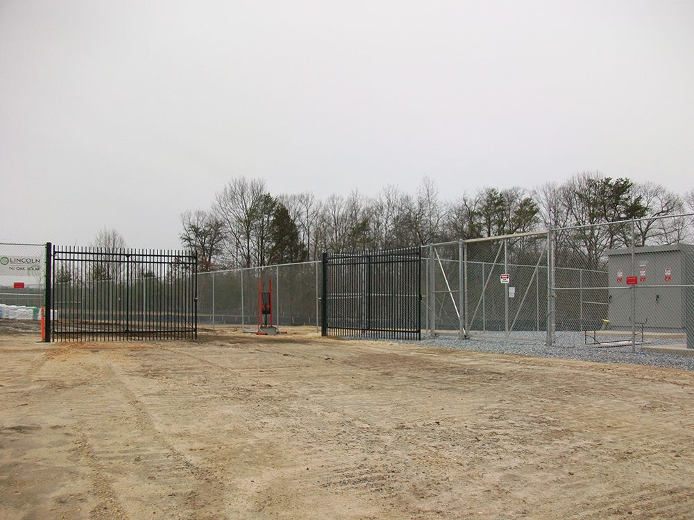 Fenced industrial area with gates and electrical equipment outdoors. Trees in the background, overcast day.