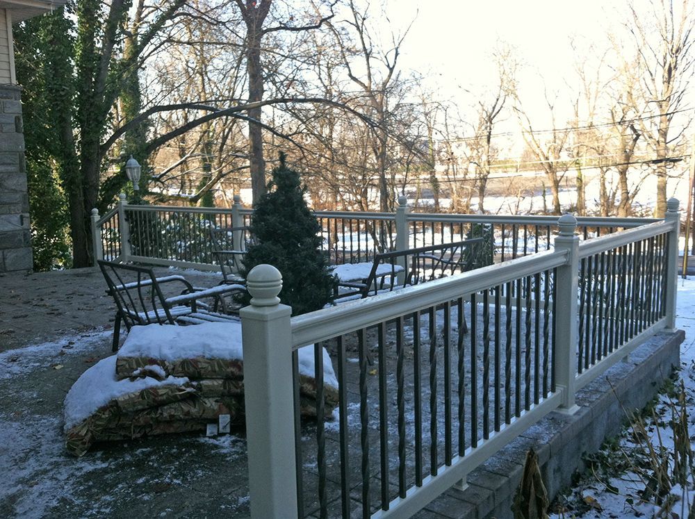 Snowy patio with black railing and furniture, overlooking a lake with trees.