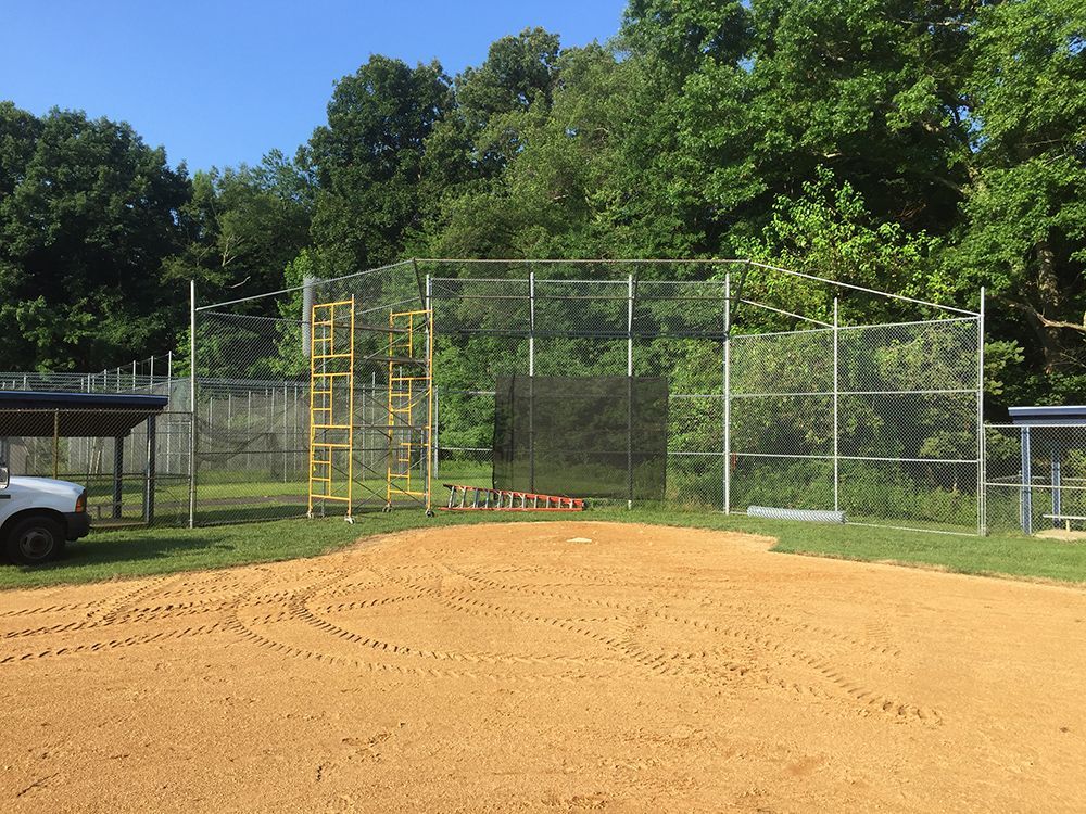 A baseball batting cage in a grassy area, with a chain link fence, trees, and dirt field.
