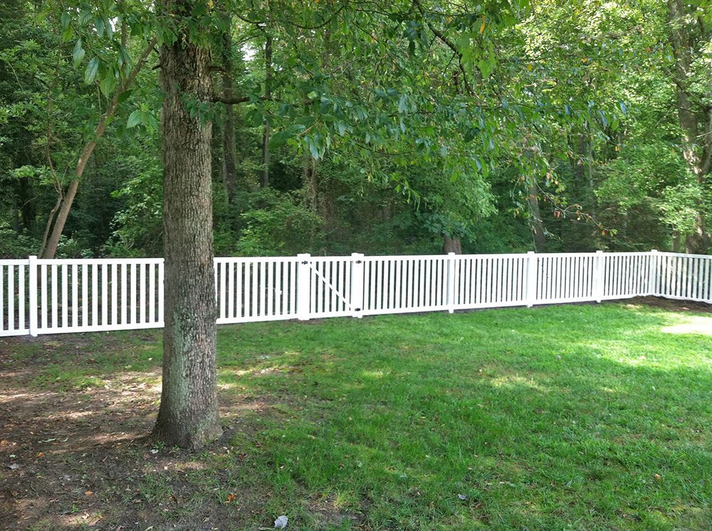 White picket fence enclosing a grassy yard near a tree, with a wooded background.