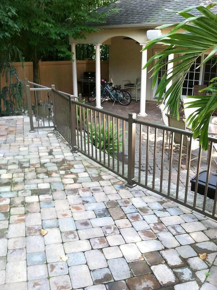 Brown metal fence borders a brick patio, leading to a covered structure with a bike inside.