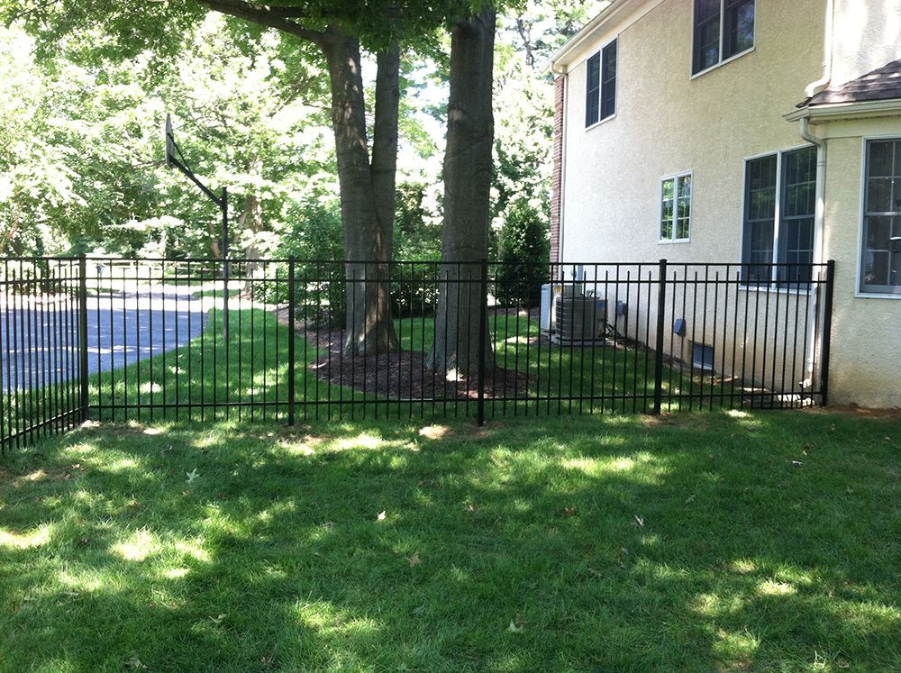 Black metal fence surrounds a grassy yard near a tree and a beige house.