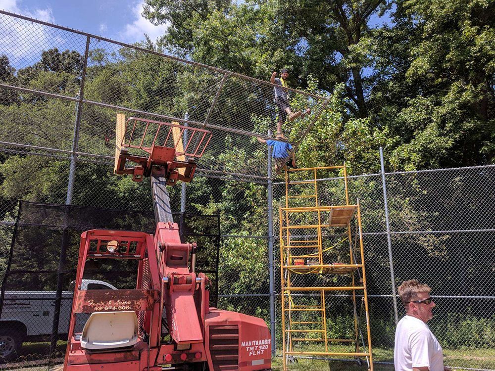 Two people climb a fence, aided by a lift and scaffolding. A person watches. Sunny, outdoor setting.