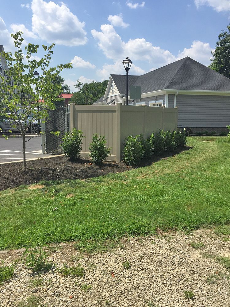 Tan vinyl fence with shrubs, small tree, and grassy area under a blue sky.
