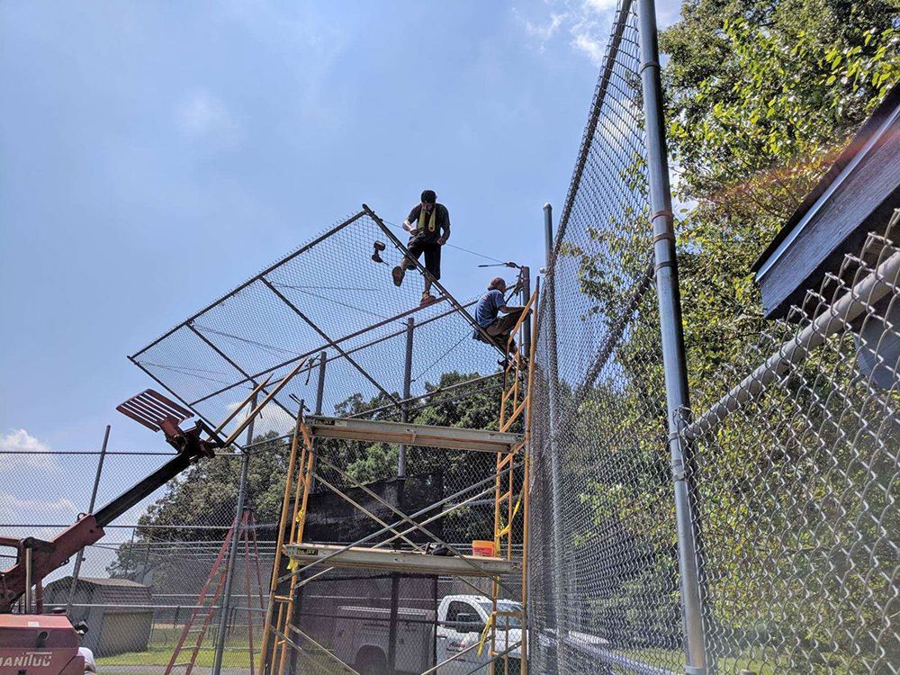 Two workers installing chain-link fence on scaffolding and a lift, under a blue sky, outside.