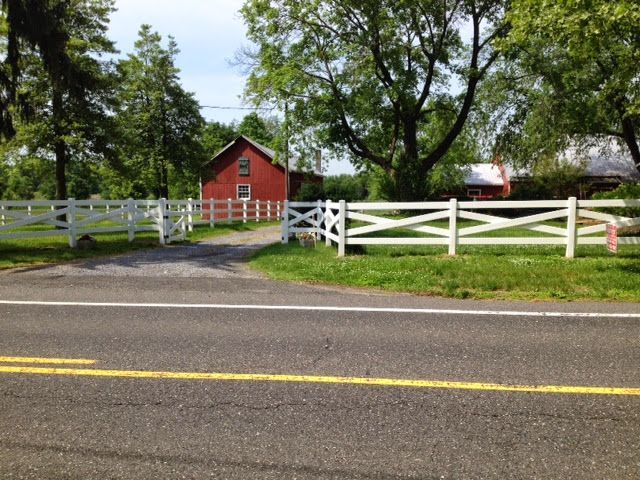 Red barn behind a white fence, driveway, and paved road; trees in background.