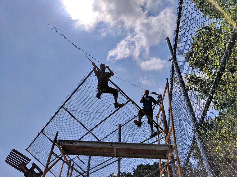 Two people on a climbing structure, reaching for ropes overhead, against a bright sky.