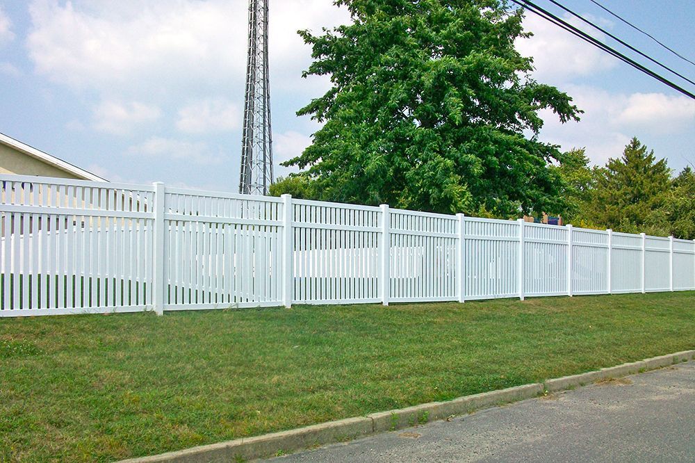 White picket fence in front of a green lawn and trees, with a building and utility pole in the background.