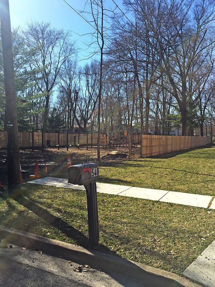 A front yard with a fence, trees, and a mailbox. Green grass and blue sky are visible.