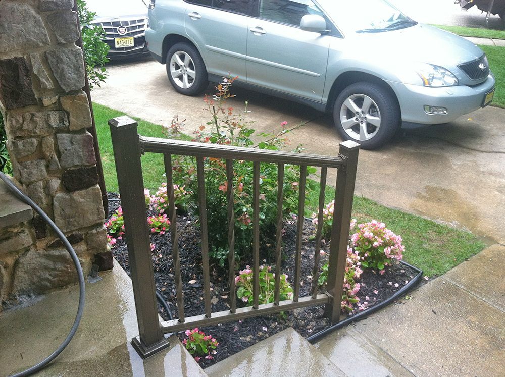 Brown railing in front of a flowerbed and driveway, next to a stone pillar, with a silver car in the background.