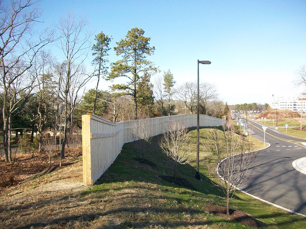 Wooden fence atop a grassy hill next to a curved road and trees on a sunny day.