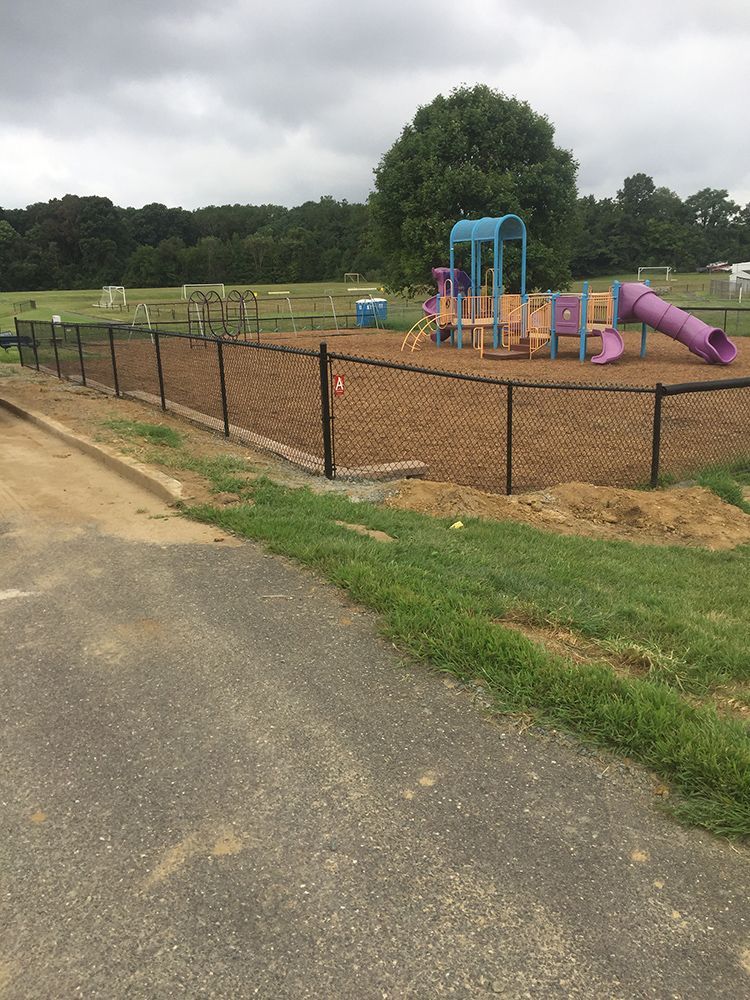 Playground with purple slide, blue climbing structure, fenced in, set in a grassy area with trees and a cloudy sky.
