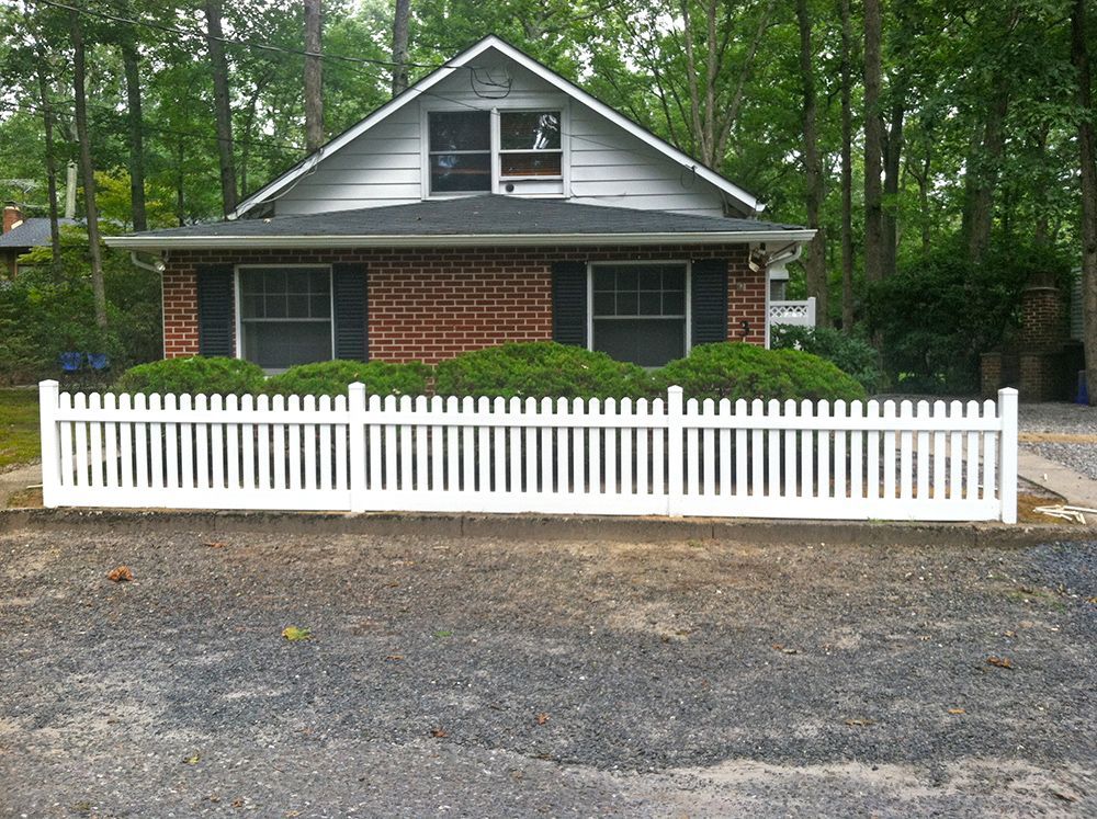 White picket fence in front of a brick house with a dark roof and green bushes.