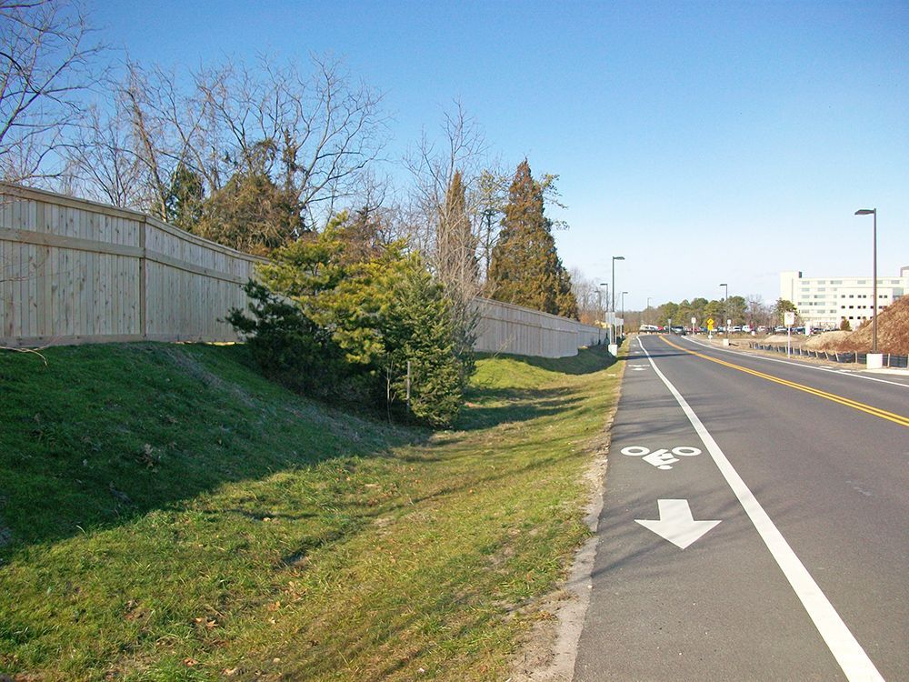 Roadside view with bike lane, grass embankment, wooden fence, and trees under a clear blue sky.