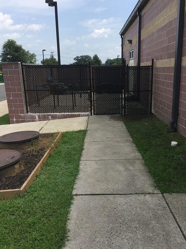 Concrete walkway leading to a black metal gate and brick building. Green grass borders the path.