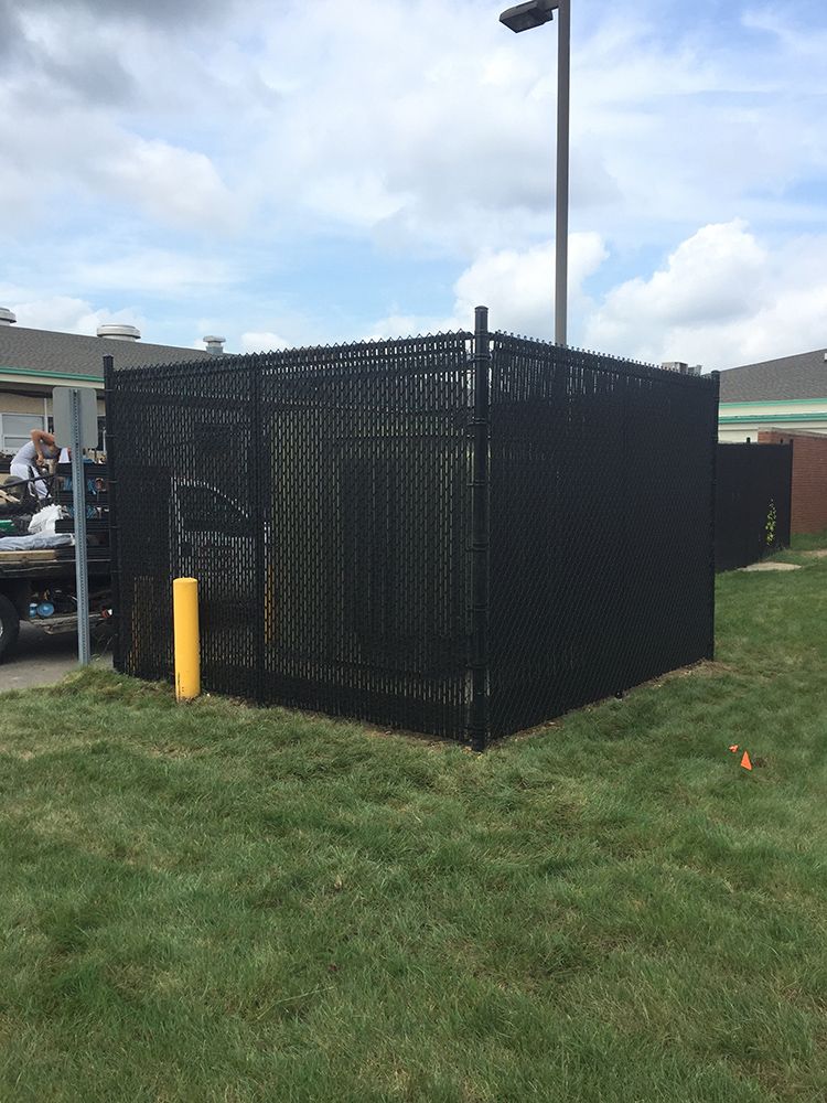 Black chain-link fence enclosure in a grassy area, possibly for machinery. A yellow post is near the fence.