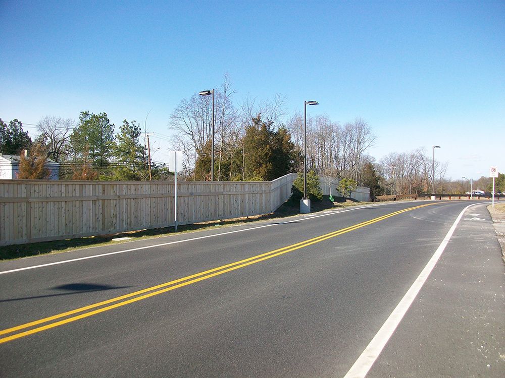 Asphalt road with yellow lines, a wooden fence, trees, and streetlights under a blue sky.