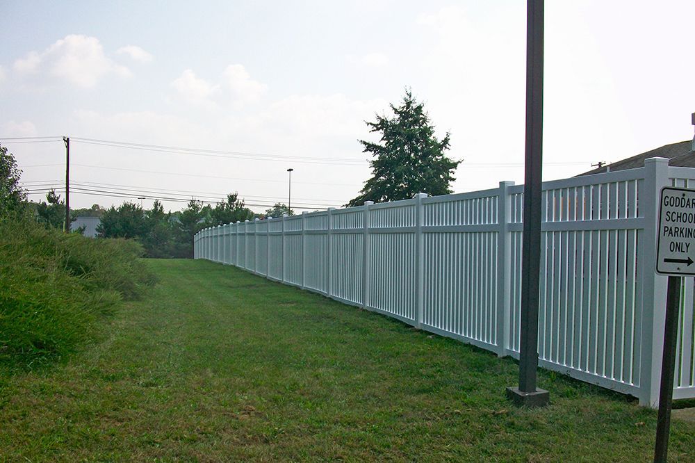 White vinyl fence along a grassy area, separating it from trees and a building.