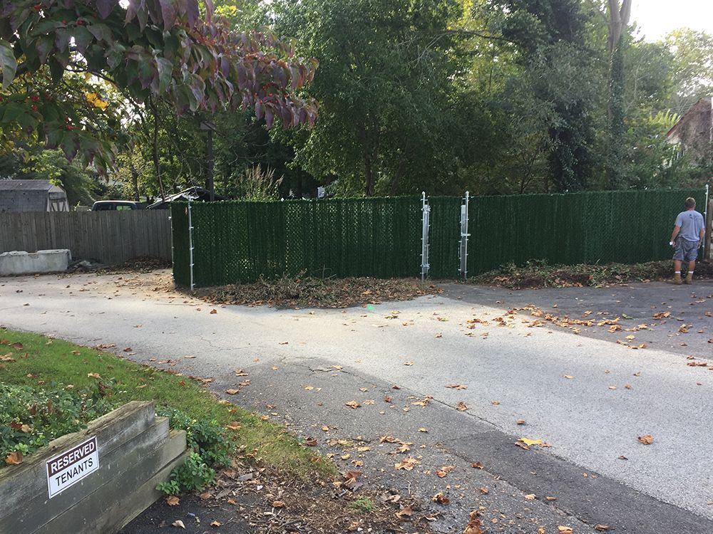 Green fence along a driveway with a person standing near it, autumn leaves scattered on the ground.