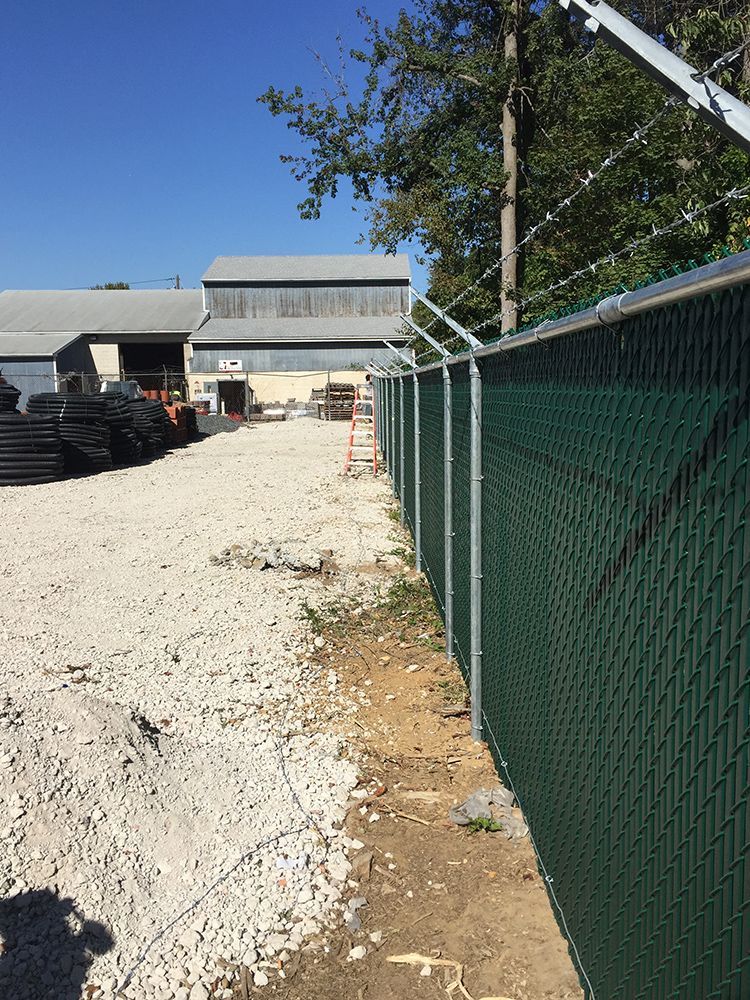 Green chain link fence with razor wire, gravel ground, industrial buildings in background.