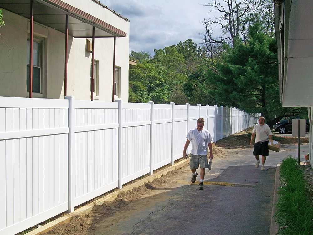 Two men walk down a paved alley next to a white fence. Buildings and trees are visible.
