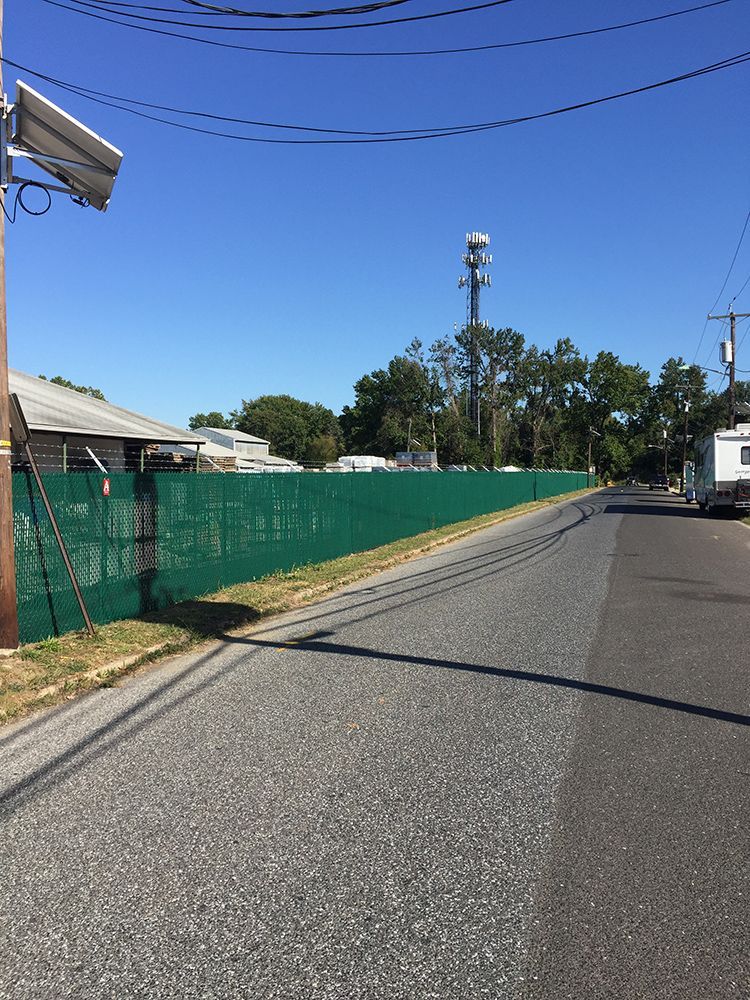 Green fence beside a paved road with a cell tower visible in the distance under a clear blue sky.