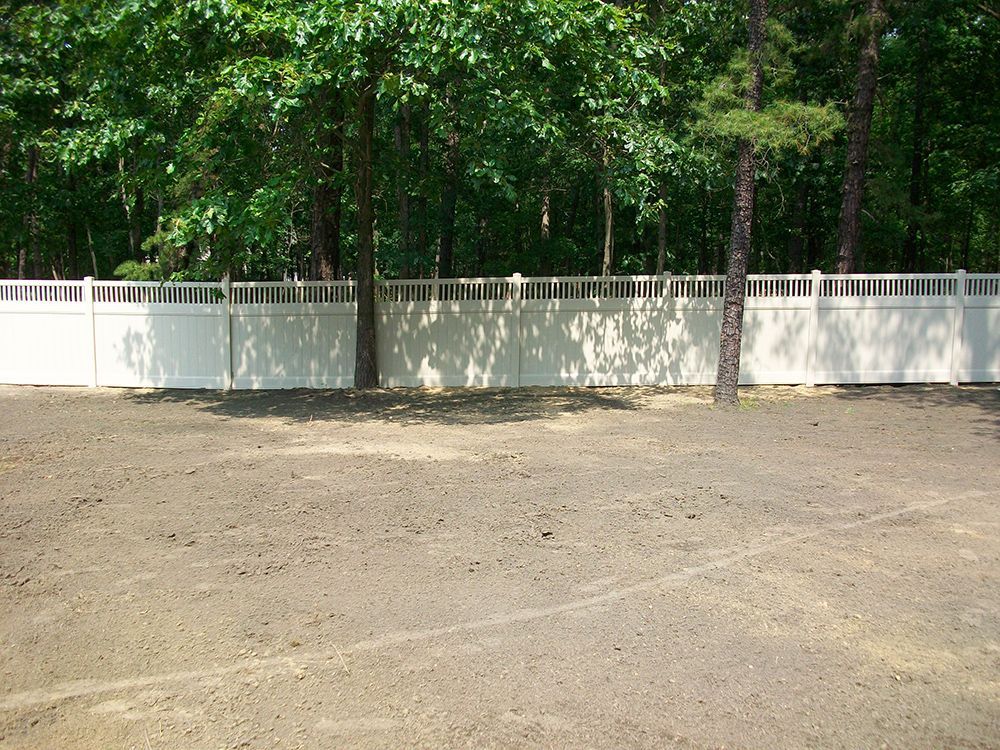 White fence in front of a forest, casting shadows on a gravel surface.