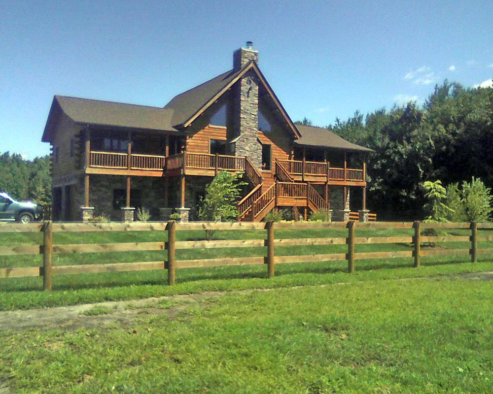 Wooden cabin with stone chimney and wrap-around porch, behind a wooden fence, on a grassy field.