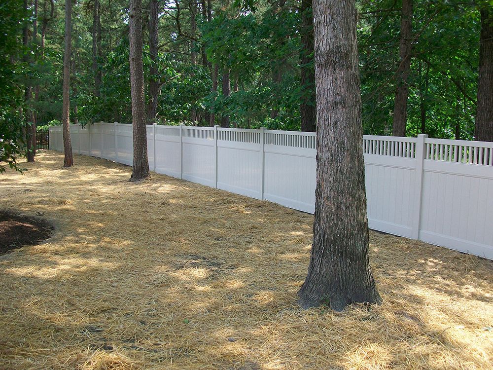 White fence in a wooded area; brown ground cover; trees along the perimeter.