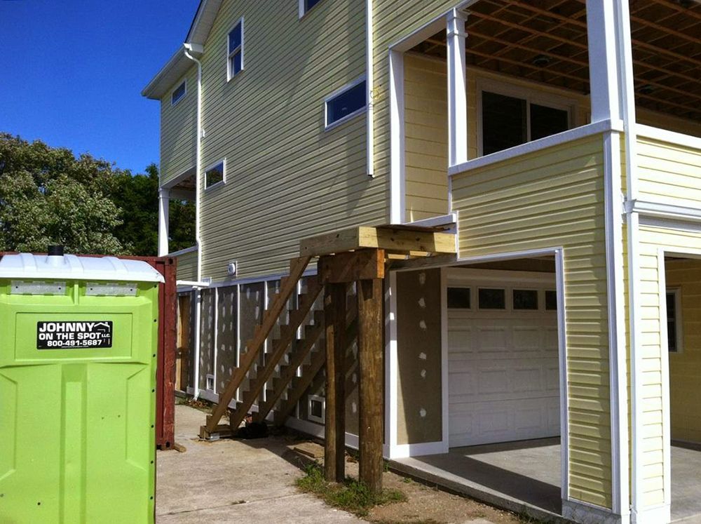 Green porta-potty next to a house with wooden stairs leading to a deck over a garage.